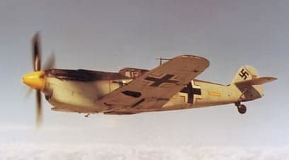 A Messerschmitt Bf 109 aircraft in flight, with a yellow nose and camouflage paint scheme.