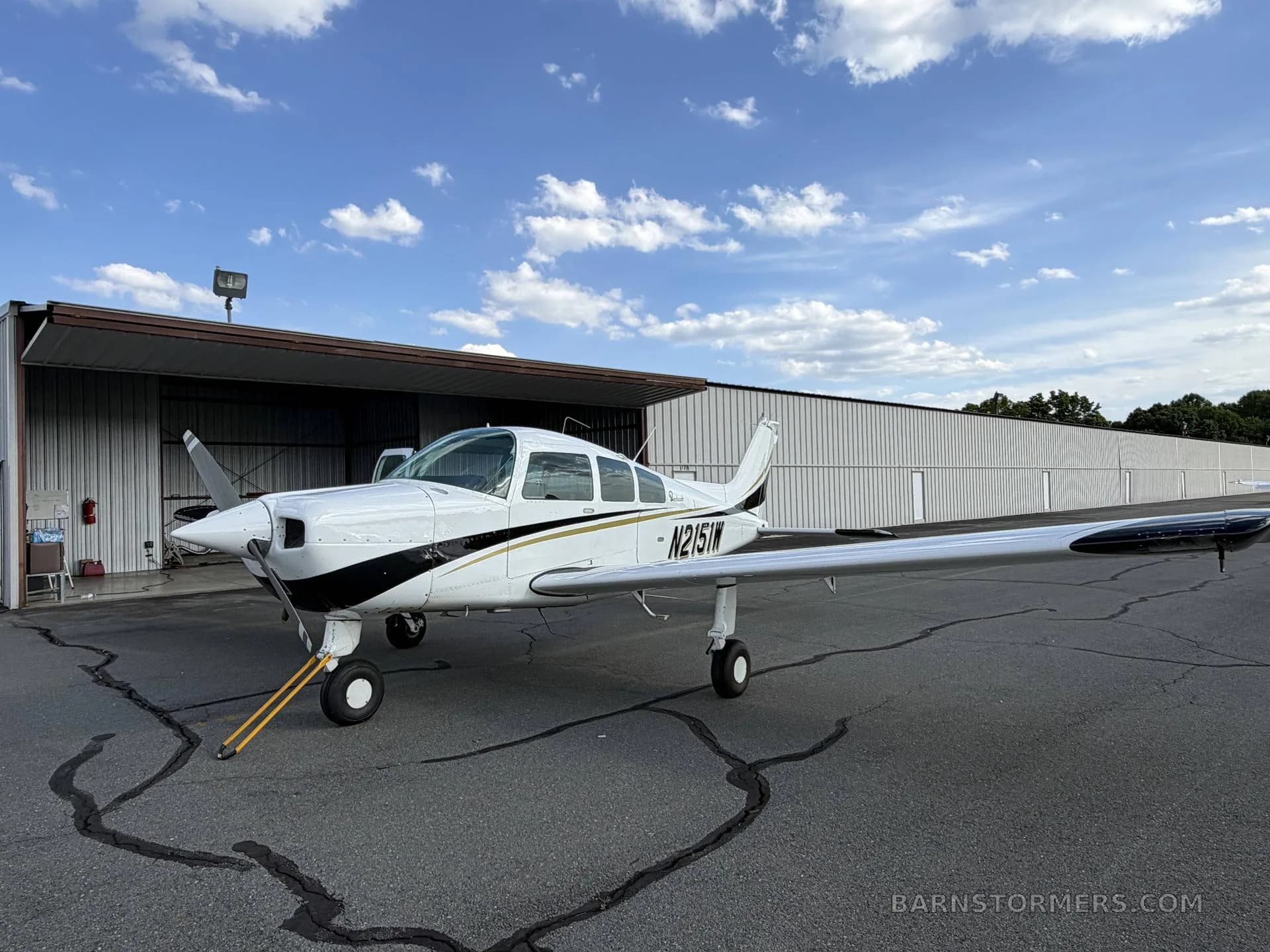 A white and black single-engine aircraft, N2151W, with gold stripes, parked on an asphalt ramp in front of a hangar under a blue sky.