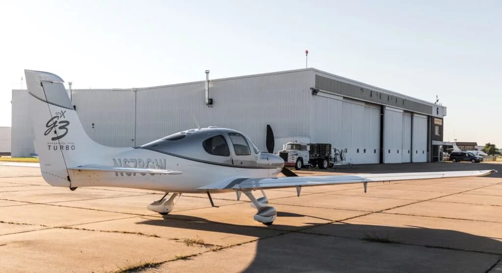 A rear quarter view of a silver and white Cirrus SR22 G3 Turbo aircraft (N678GW) parked in front of a large hangar.