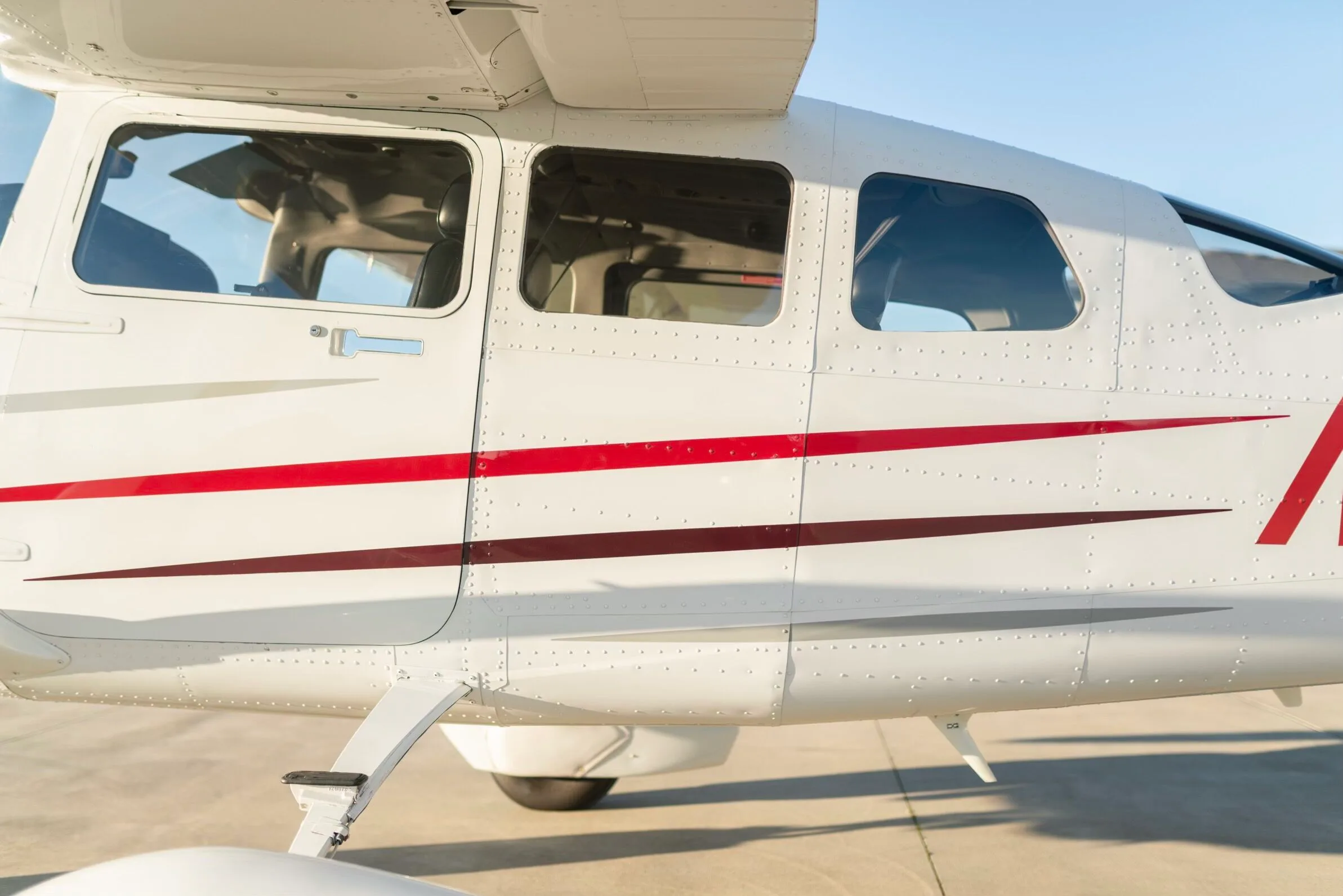 A close-up of the white fuselage of a Cessna aircraft, showing the windows and red and dark red accent stripes.