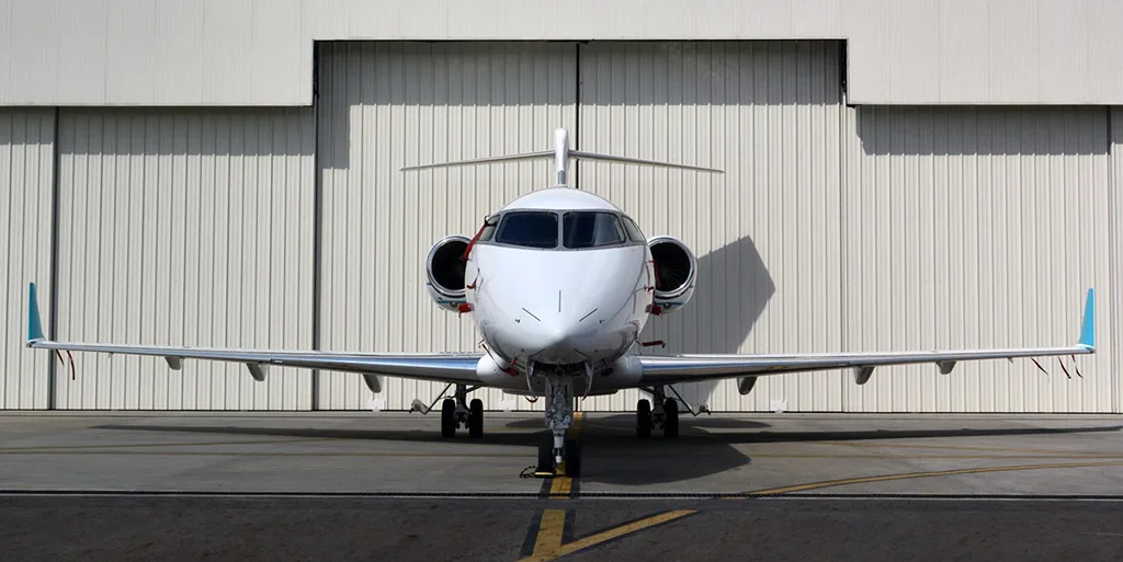 A white business jet with blue and teal stripes is viewed head-on, parked on a tarmac in front of a hangar.