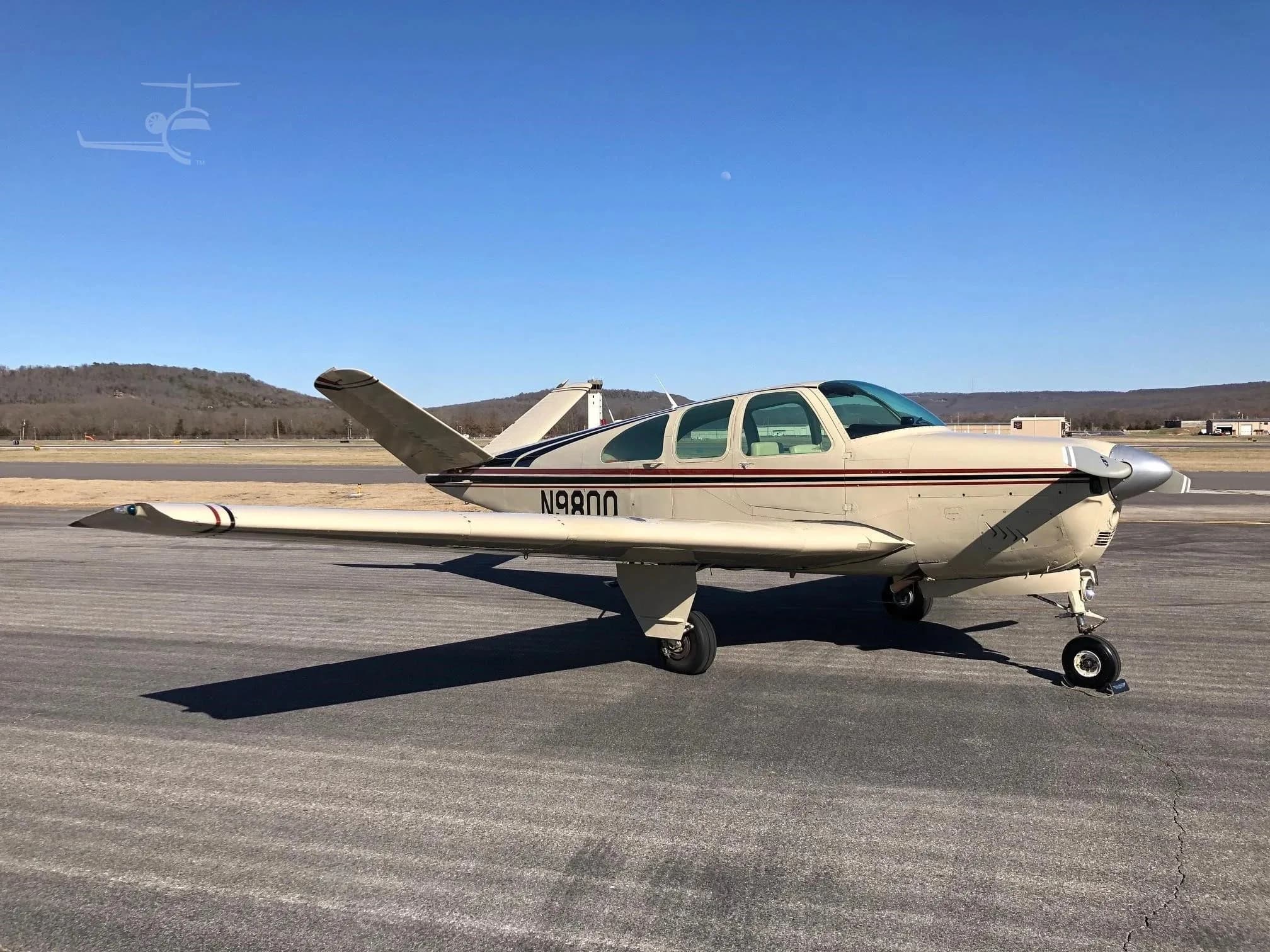 A beige Beechcraft Bonanza N980Q with red and black stripes parked on an asphalt runway under a clear sky.