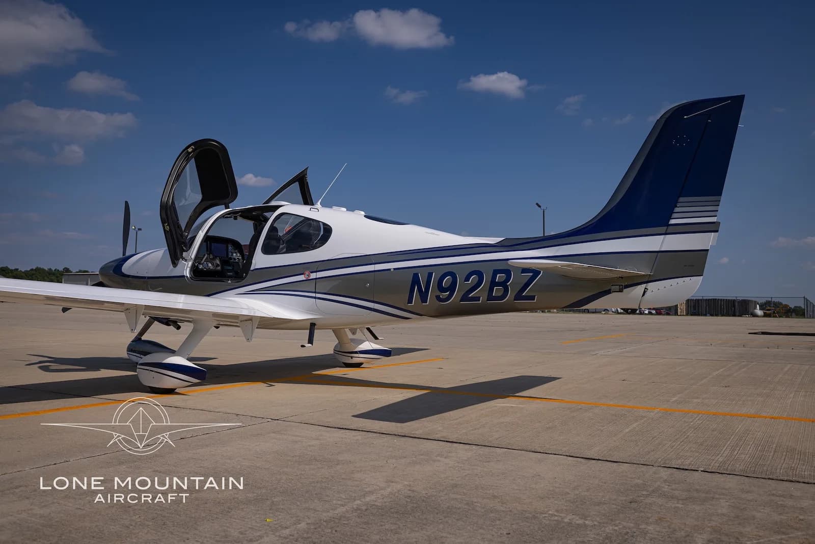 A three-quarter rear view of the white and silver Cirrus SR22 aircraft N92BZ with blue accents, parked on an airfield with its doors open.