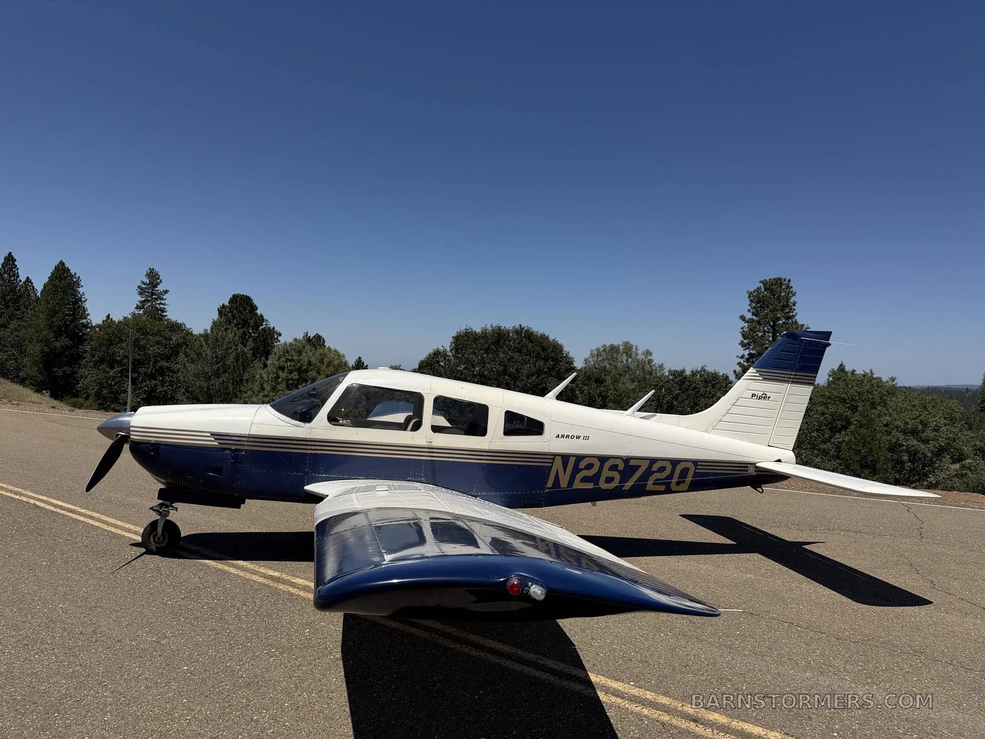 A blue and white Piper Arrow III (N26720) parked on an asphalt surface with trees in the background under a clear blue sky.