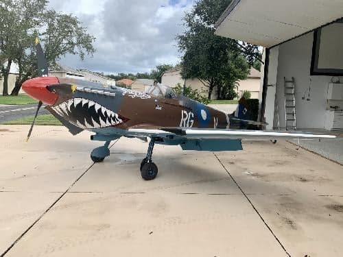 A P-40 Warhawk replica aircraft with shark mouth paint scheme is parked on a concrete pad.