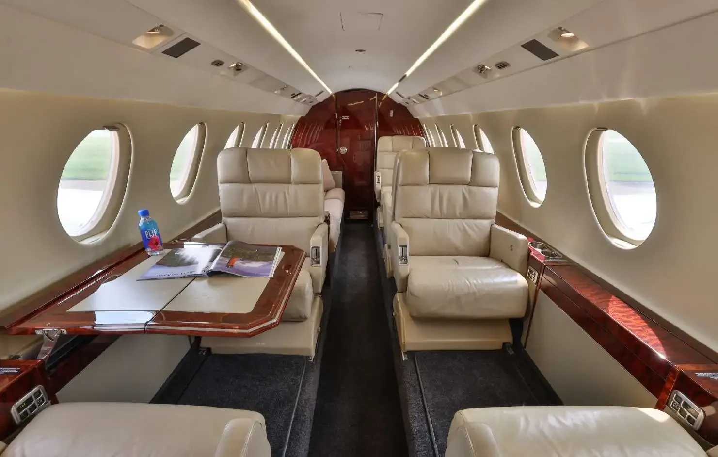 The luxurious interior cabin of a private jet, featuring beige leather seats, a wooden table, and large windows.