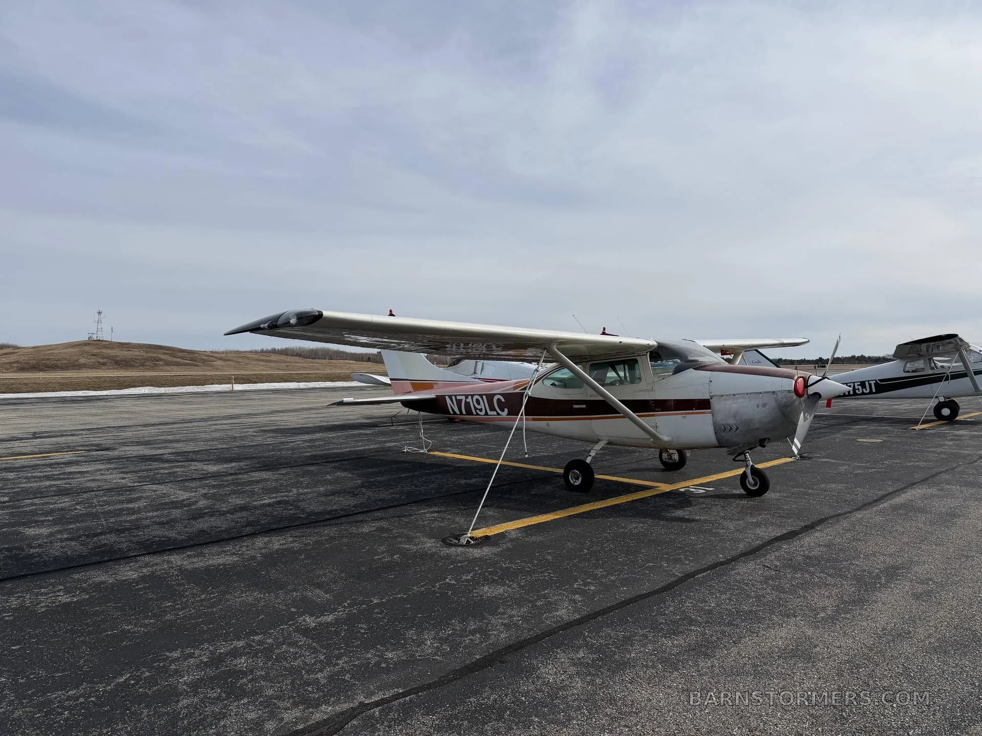 A white and brown Cessna-style aircraft with registration N719LC is parked on an asphalt ramp under a cloudy sky.