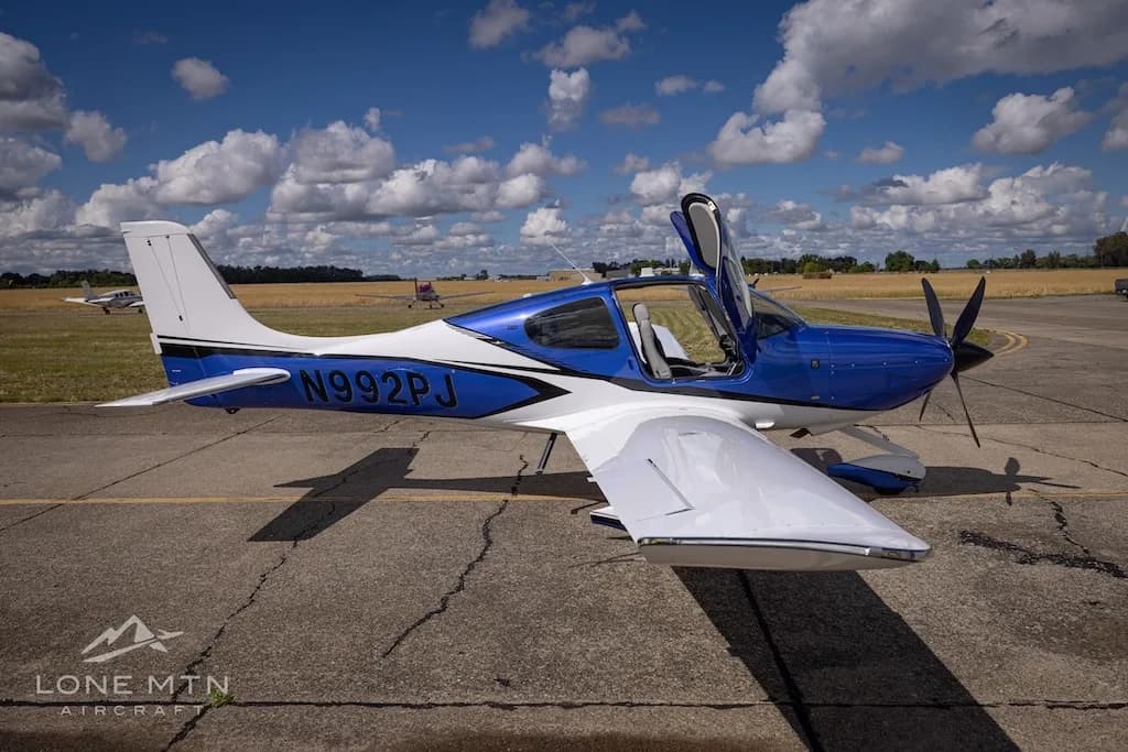 A blue and white Cirrus SR22 aircraft with its doors open, parked on an airfield with other aircraft in the distance.