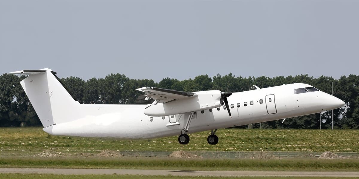 A white turboprop aircraft (likely a Dash 8) with landing gear extended, just above the runway, with green trees in the background.