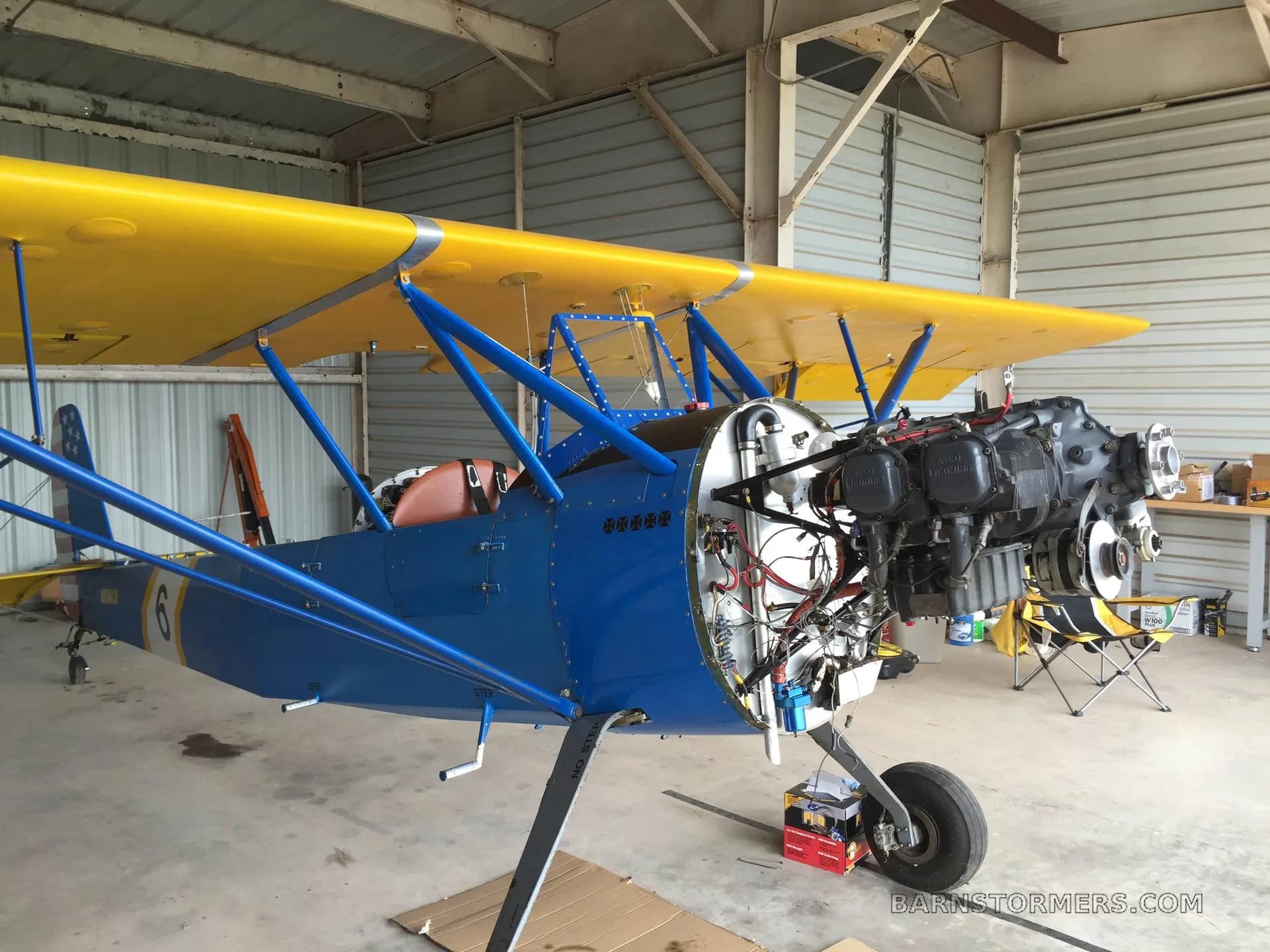 A blue and yellow biplane in a hangar with its engine cowling removed, revealing the engine components.