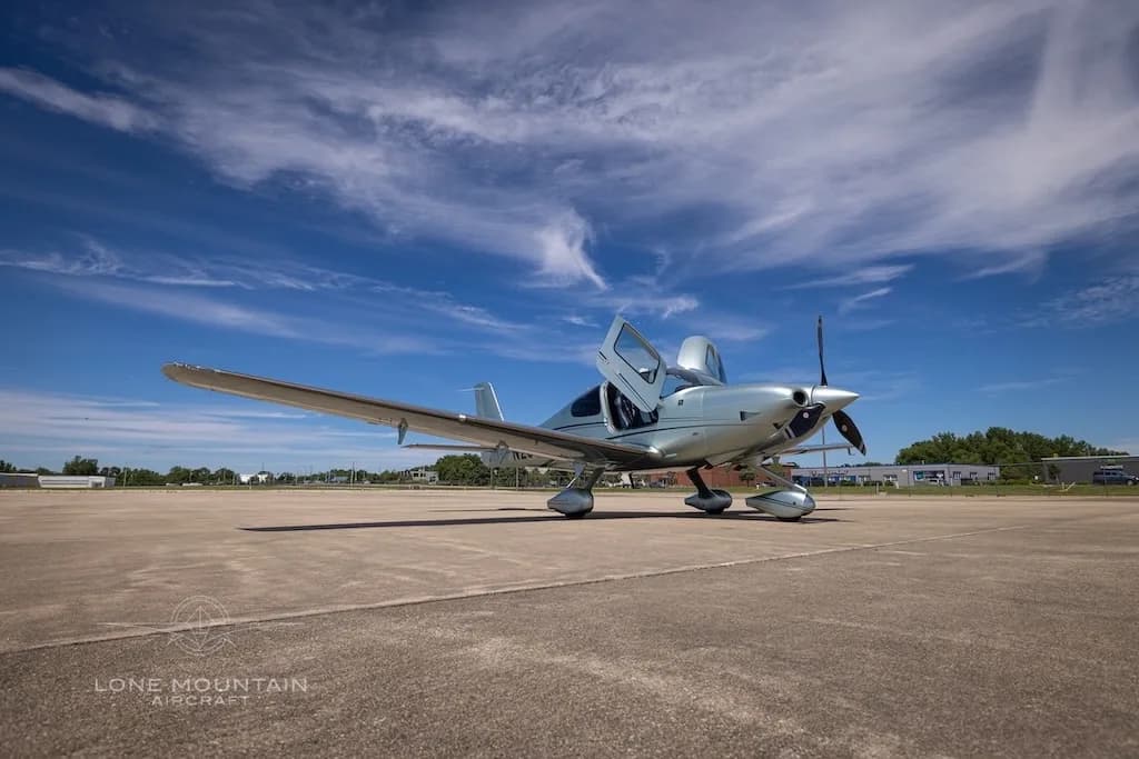A wide shot of a silver Cirrus SR22 aircraft (N234PY) with open doors on a tarmac under a bright blue sky.