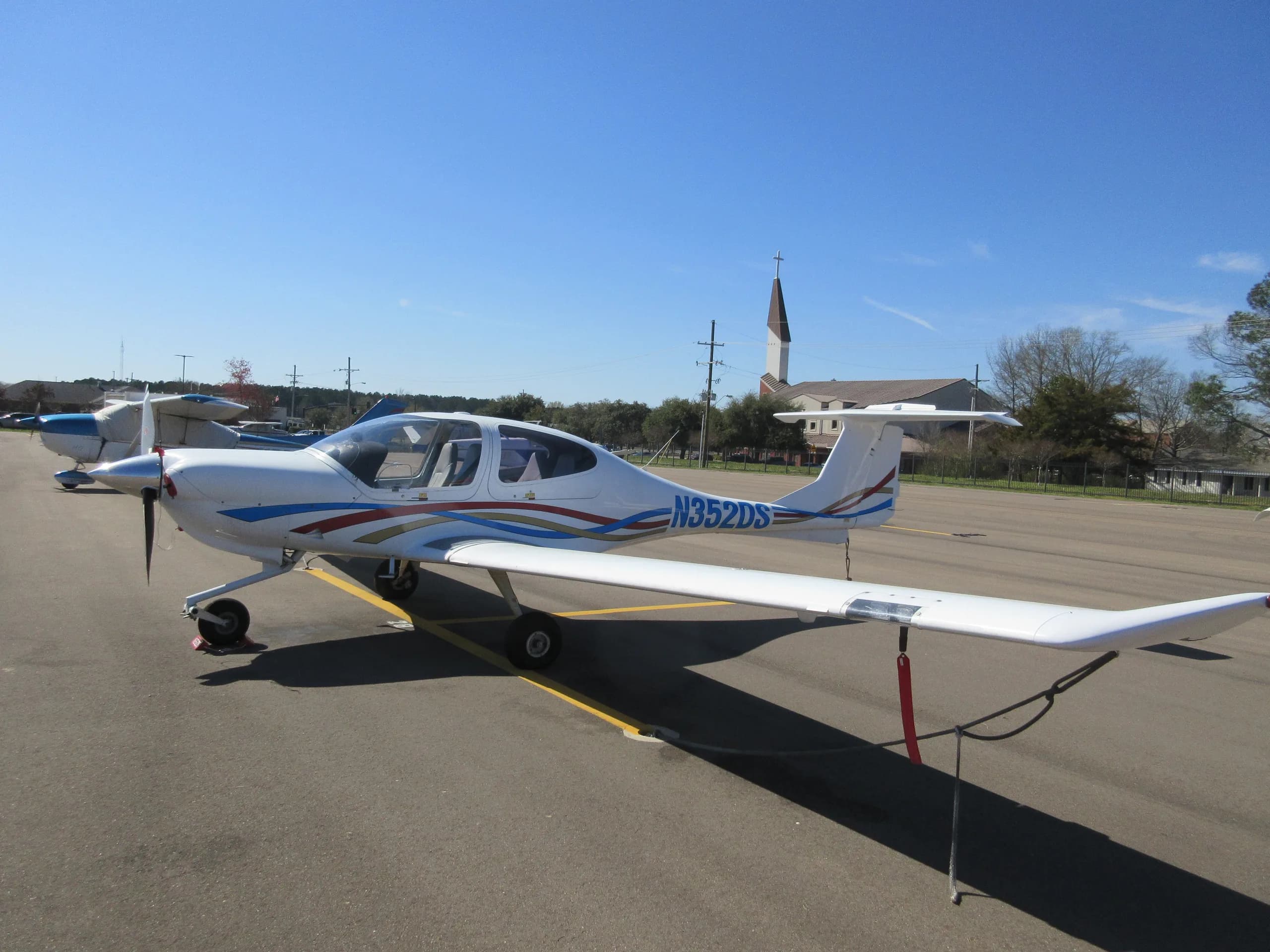 A white Diamond DA40 aircraft with blue, red, and gold stripes, registration N352DS, parked on an airport tarmac with a church steeple in the background.