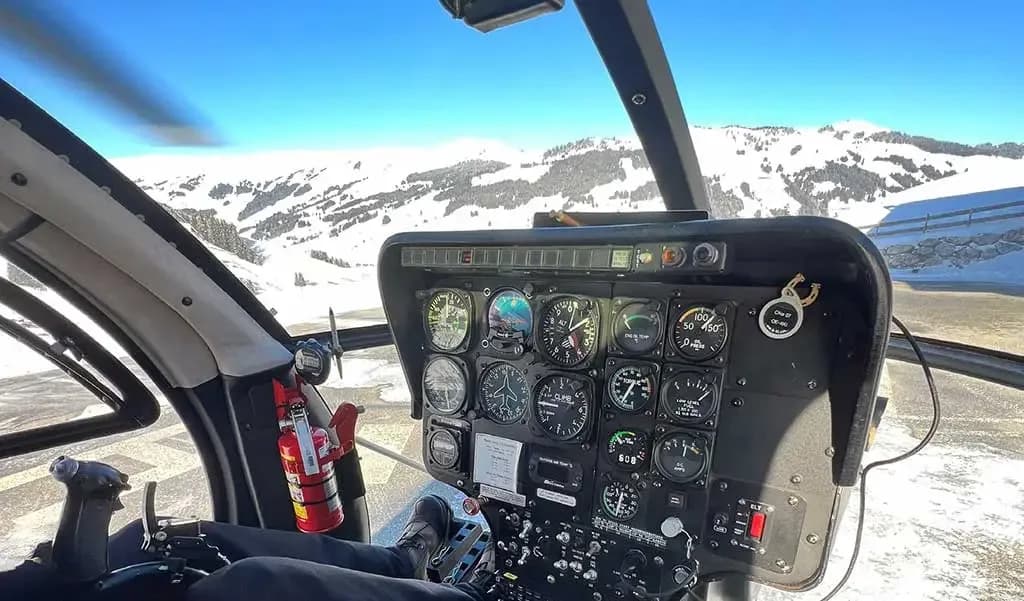 The cockpit of a helicopter shows the instrument panel and a view of snowy mountains through the windshield.