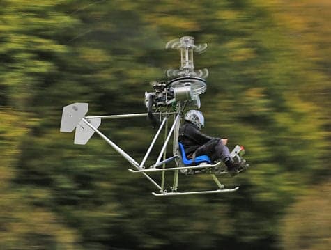 A single-seat helicopter flying in front of trees.