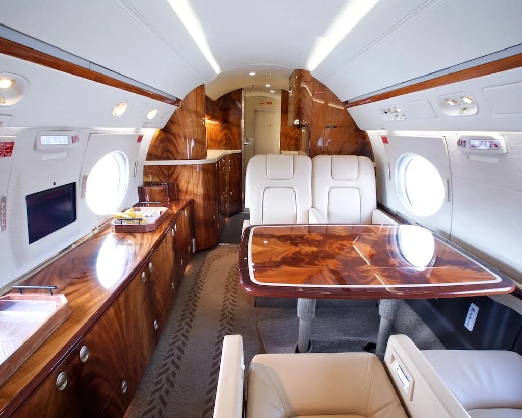 The interior of a private jet featuring a dining table, beige seats, and a galley area with polished wood cabinetry.