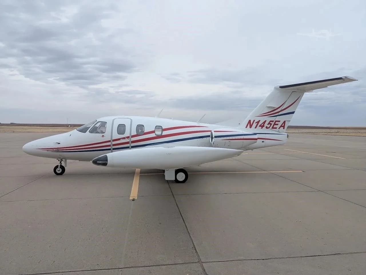 A white Eclipse jet with red and blue stripes, registration N145EA, parked on an airfield from a side view.