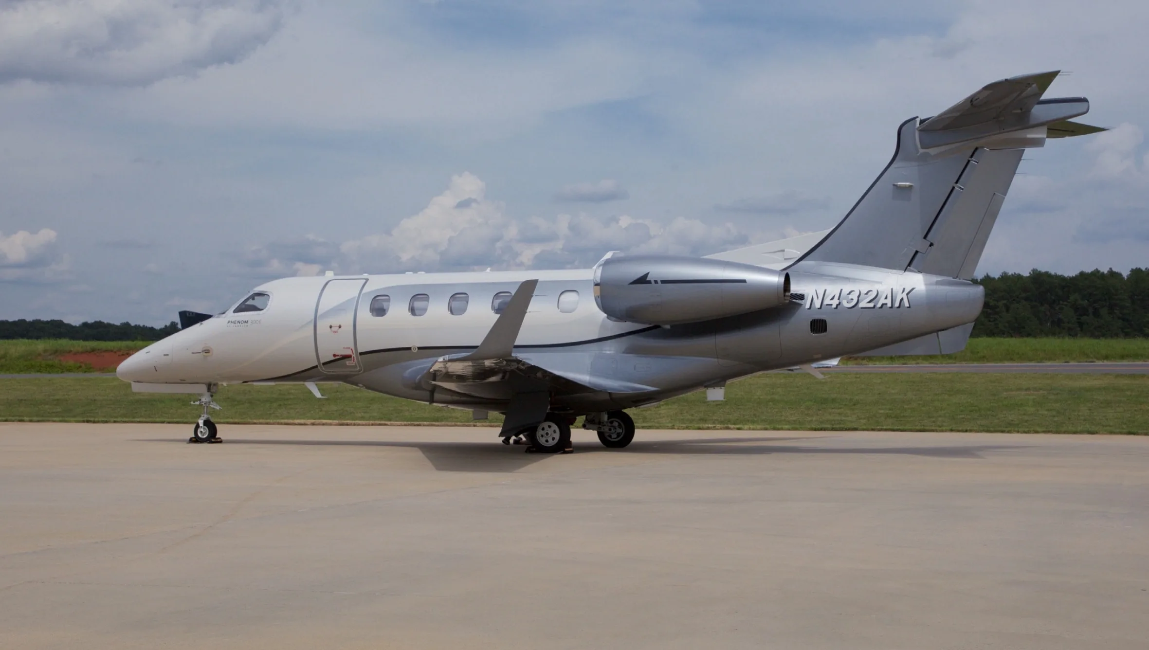 A side view of a white and grey Embraer Phenom 300E business jet, N432AK, parked on an airfield under a cloudy sky.