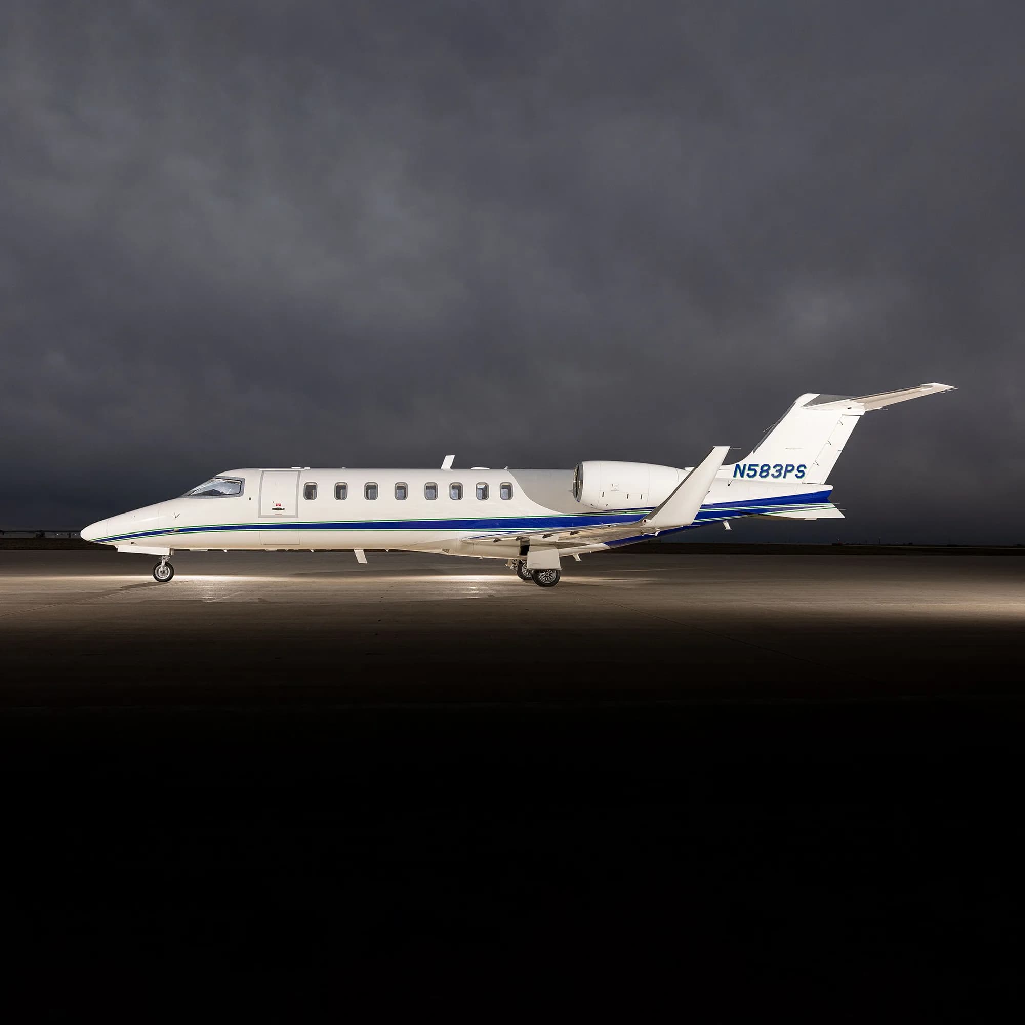 A white Learjet 75 with blue and green stripes, registration N583PS, parked on an tarmac under dramatic cloudy skies.