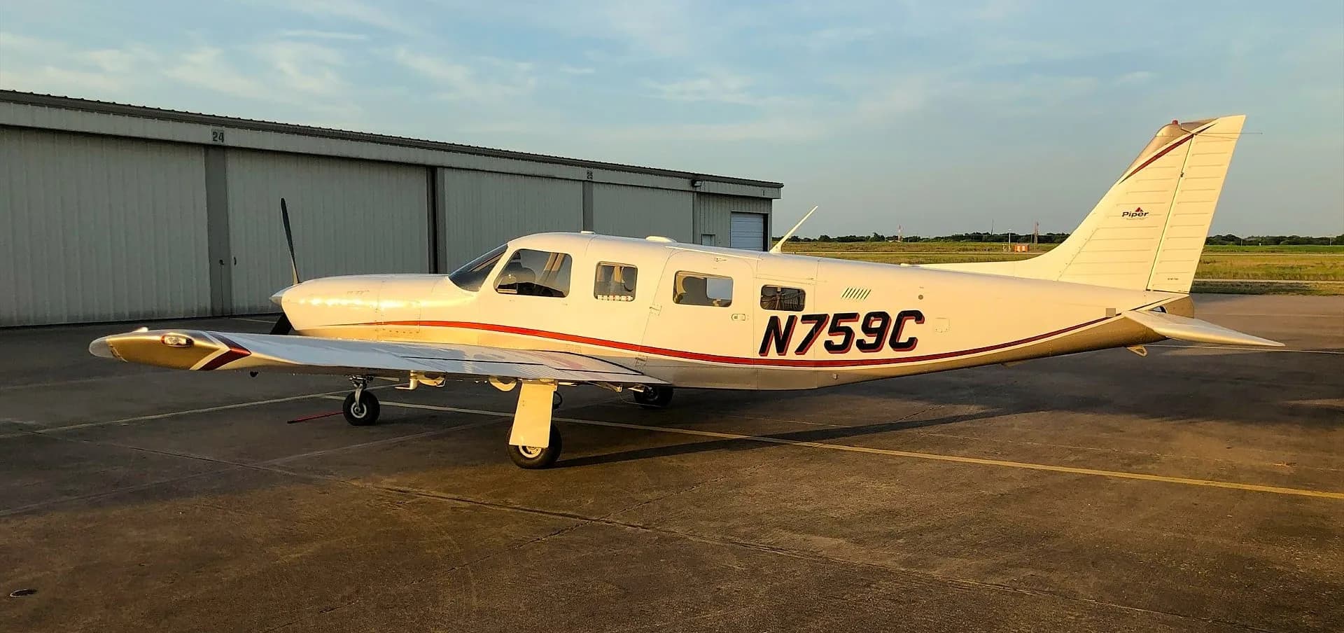 A white and tan Piper Saratoga II TC aircraft (N759C) parked on an asphalt ramp in front of a hangar at sunset.
