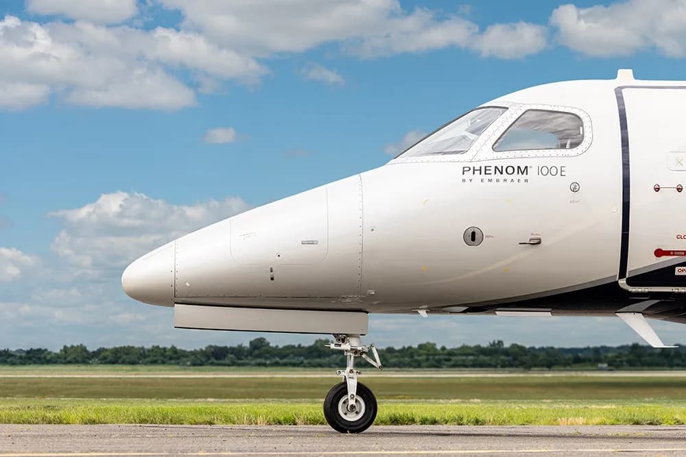 A side view of the nose and front landing gear of the white Embraer Phenom 100E business jet on an airfield.