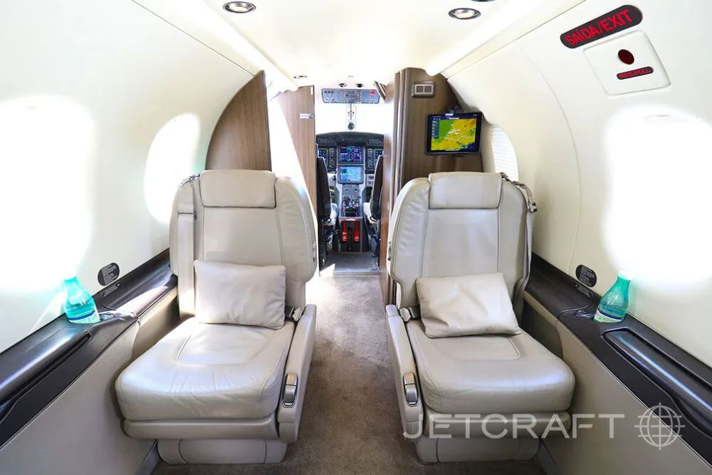 The interior cabin of a Pilatus PC-12 aircraft with light beige leather seats, looking forward towards the cockpit.