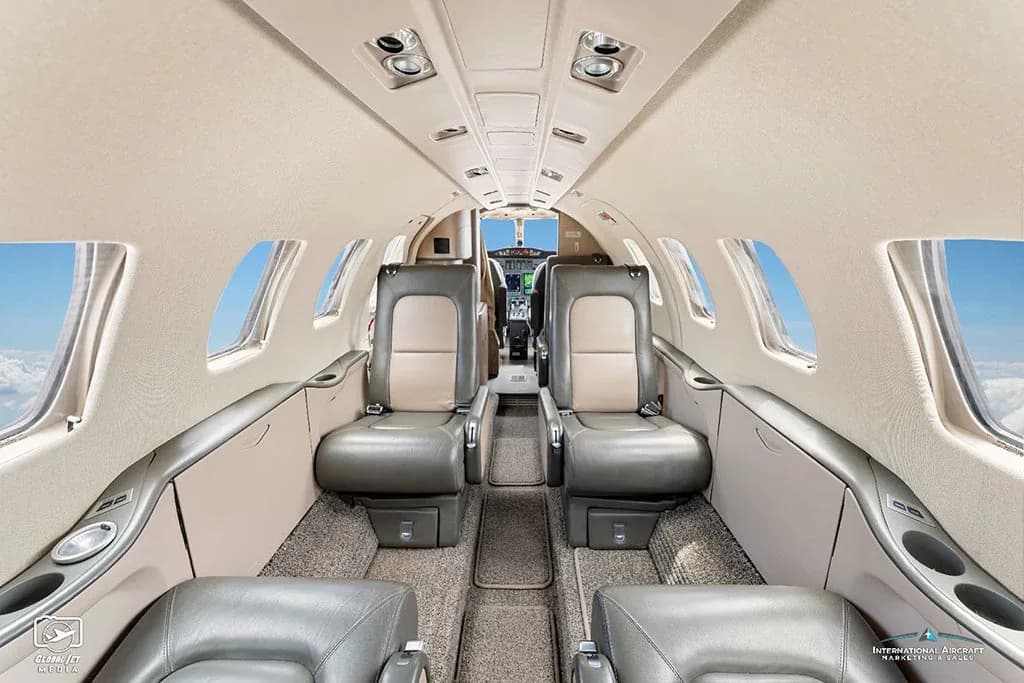 The interior cabin of a business jet features grey and tan leather seats with windows showing clouds outside.
