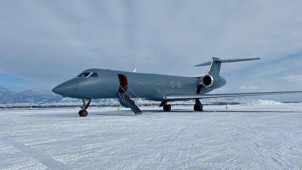 A grey Gulfstream G550 aircraft with its door open, parked on a snowy airfield with mountains in the background.
