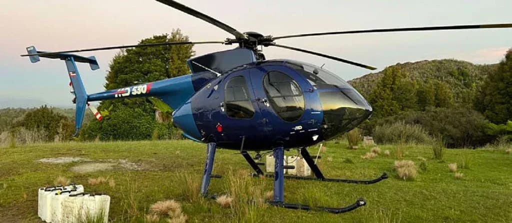 A blue and light blue MD 530 helicopter parked in a grassy field with trees and mountains under a sunset sky.