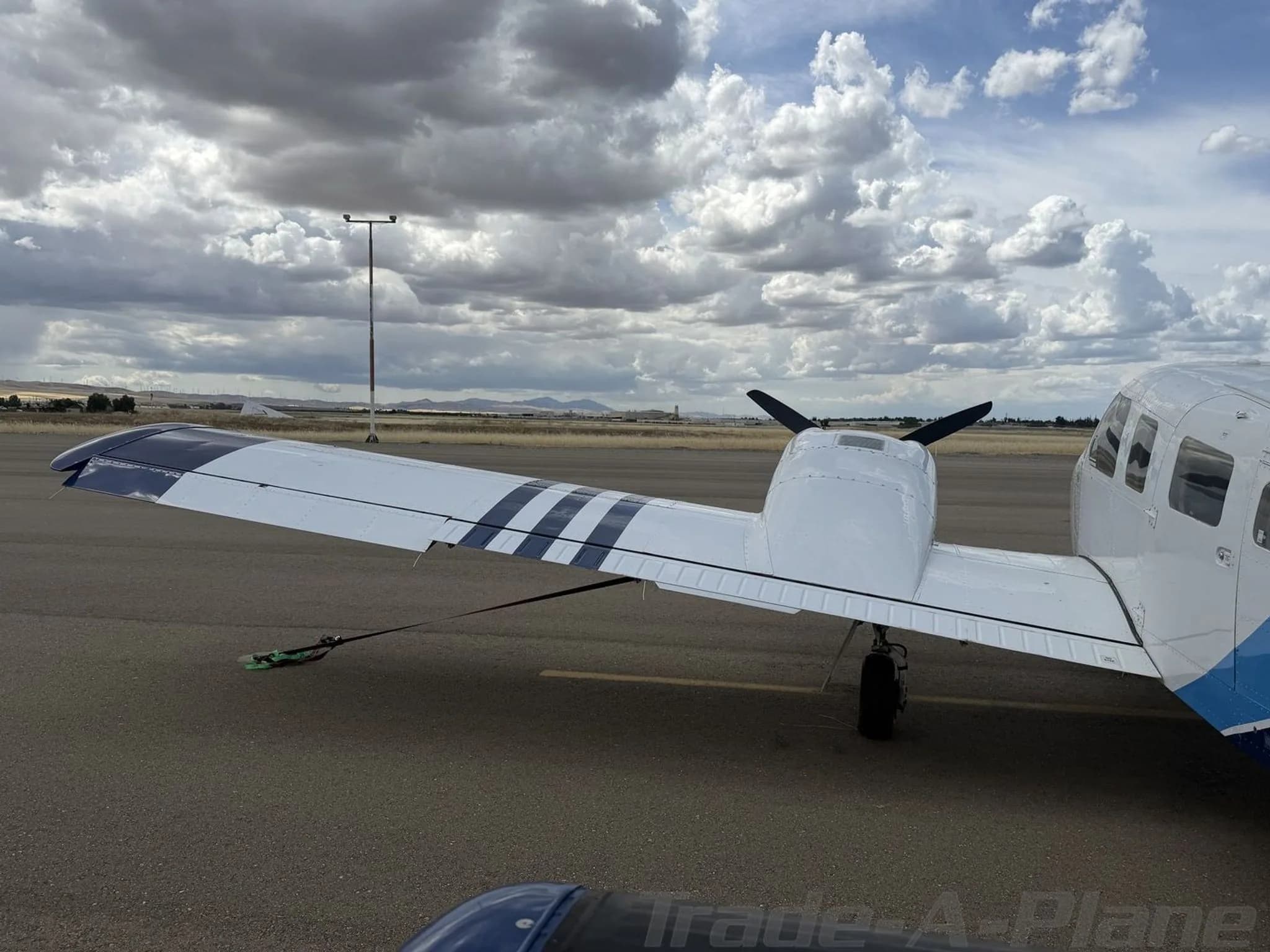 A rear-left view of the wing and fuselage of a white Piper Seneca II aircraft with blue stripes on the wing, tied down on an airport tarmac.