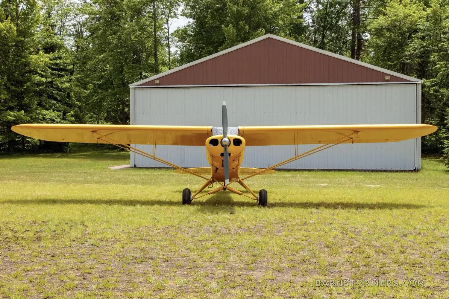 A front view of a yellow high-wing taildragger aircraft with a propeller, parked on a grassy field in front of a hangar.
