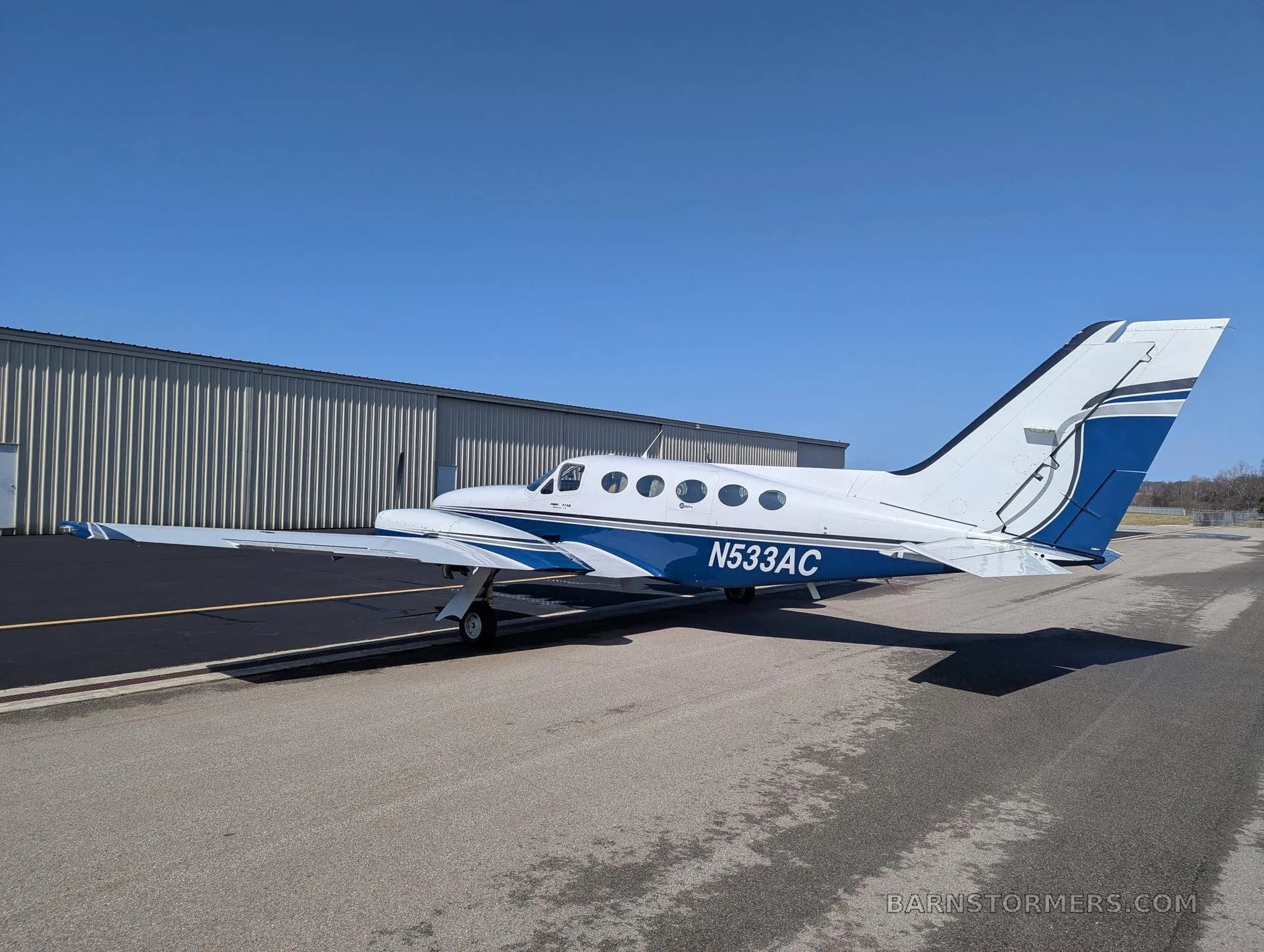 A white and blue twin-engine propeller aircraft (Cessna 414 Chancellor) with tail number N533AC parked on an asphalt ramp next to a hangar.