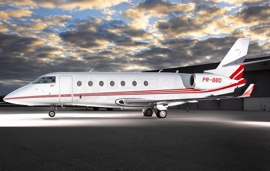 A white business jet with red stripes and registration PR-BBD is parked on a tarmac under a dramatic cloudy sky.