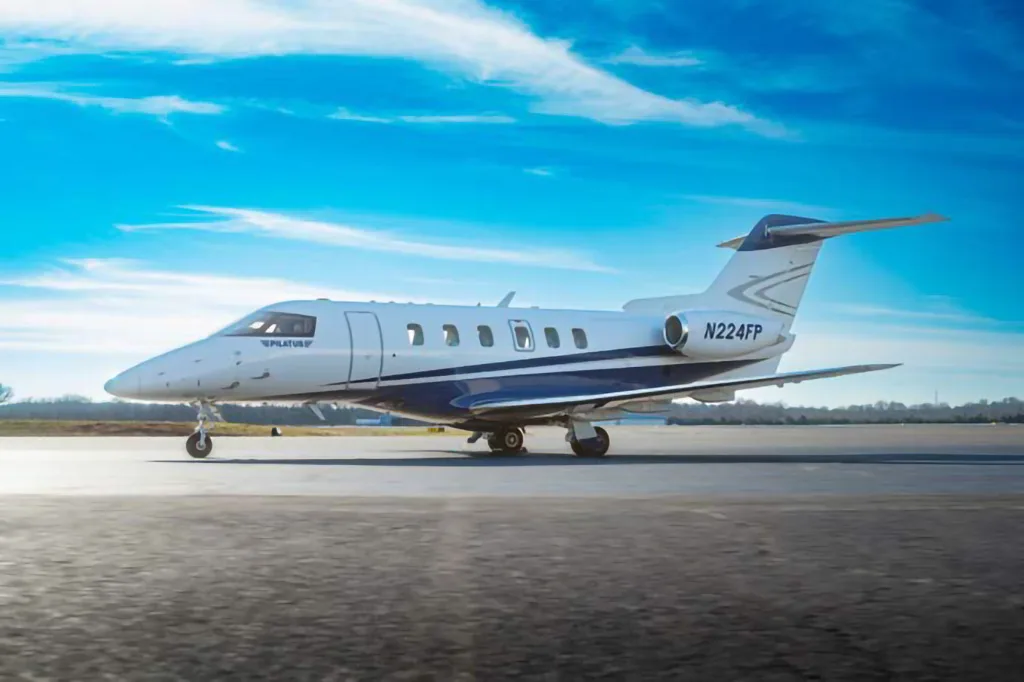A Pilatus PC-24 jet with tail number N224FP, painted white and blue, sits on an airfield under a blue sky.