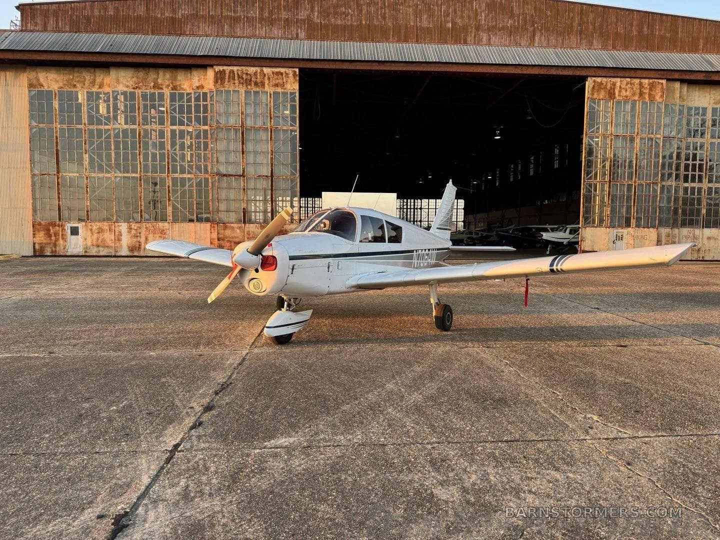 A white single-engine propeller aircraft with dark stripes parked on a concrete ramp in front of an old hangar at sunset.