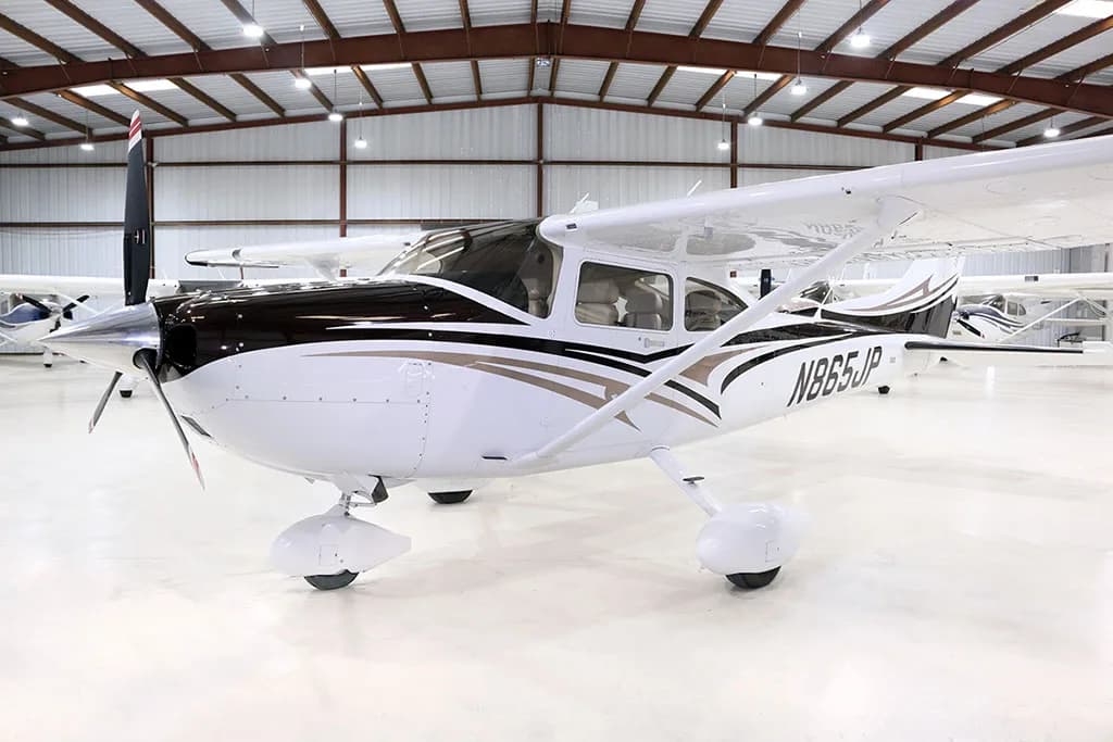 A white and black Cessna Skylane N865JP with brown accents is parked in a hangar, viewed from the front-left side.