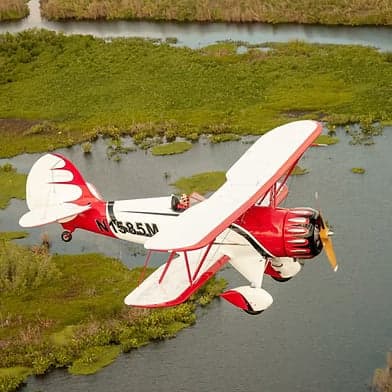 A red and white biplane flies over a swampy area.