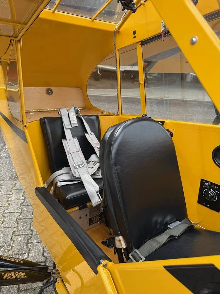 Interior view of a yellow aircraft cabin, showing two black leather seats with grey harnesses and a wooden floor.