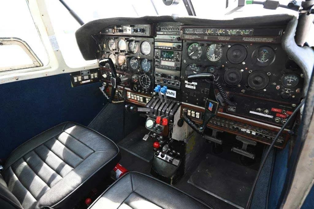 The cockpit of a twin-engine aircraft, featuring a complex instrument panel with analog gauges, switches, and throttles, and two black leather seats.
