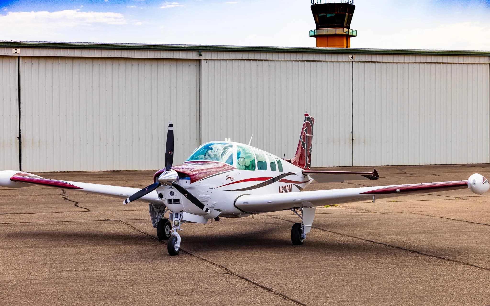 A white and maroon Beechcraft Bonanza A36 is parked on an airport tarmac in front of a hangar with an air traffic control tower in the background.
