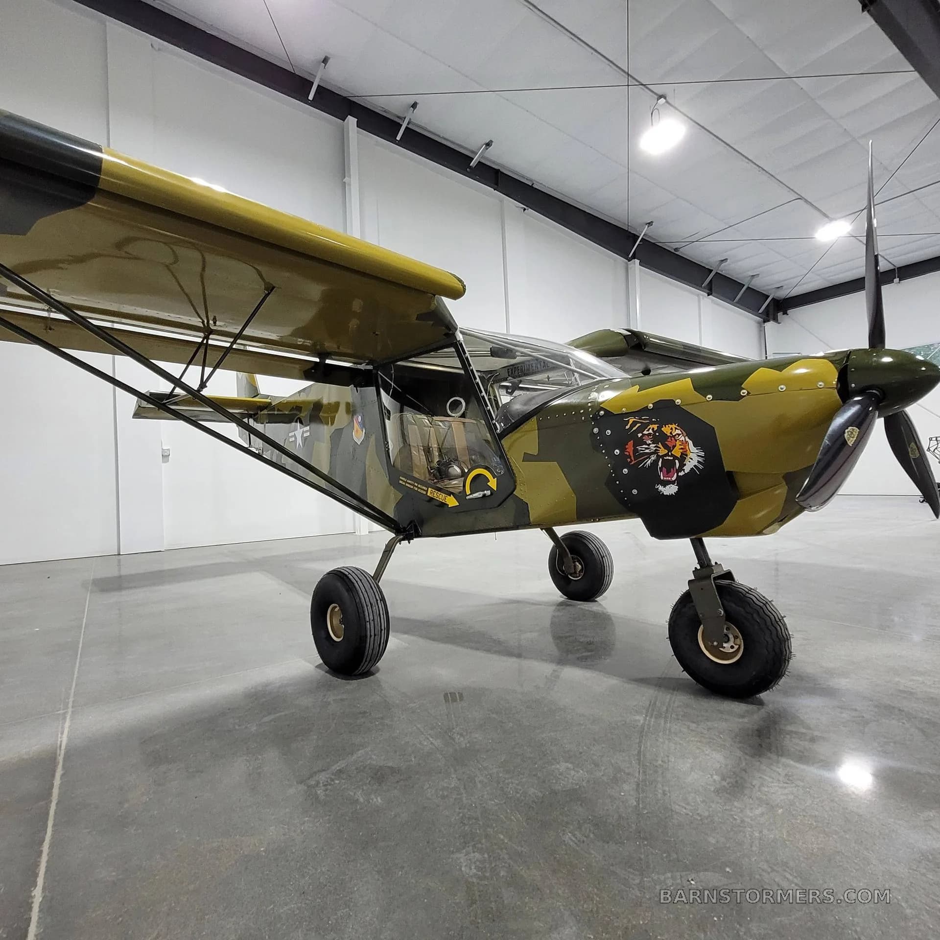A three-quarter front view of a camouflage-painted, high-wing aircraft with a tiger decal on the cowling, inside a hangar.