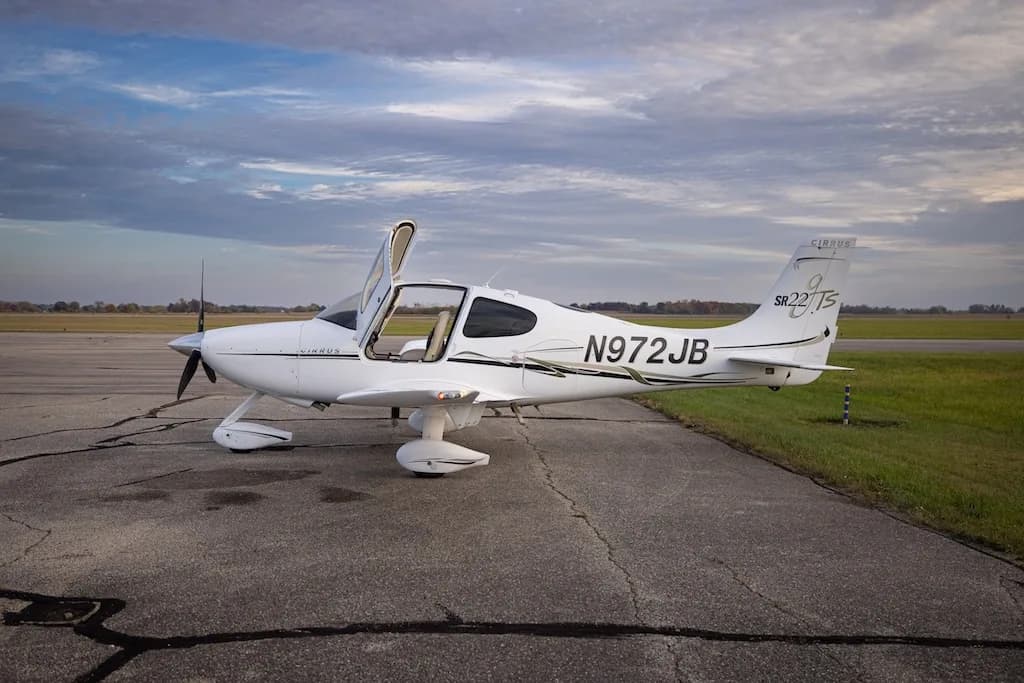 A white Cirrus SR22T aircraft, N972JB, is parked on an airfield with its doors open, viewed from the left side.