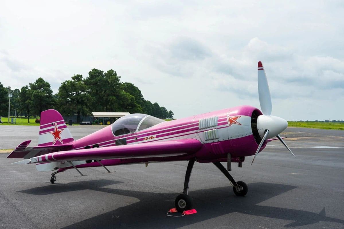 Side view of a pink and white Sukhoi Su-26 aircraft parked on the tarmac.