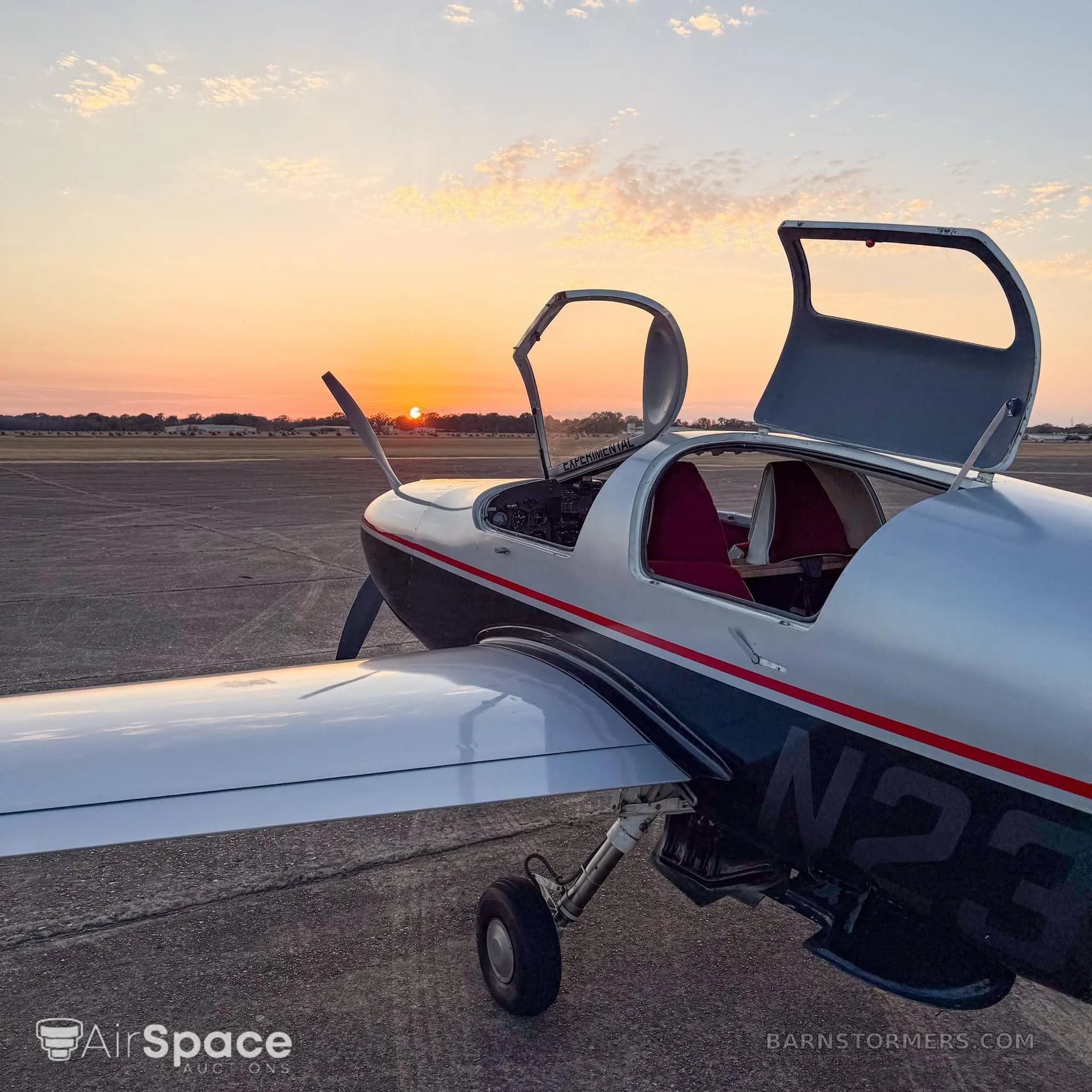 A silver and black single-engine aircraft with its canopy doors open, parked on an airfield at sunset.