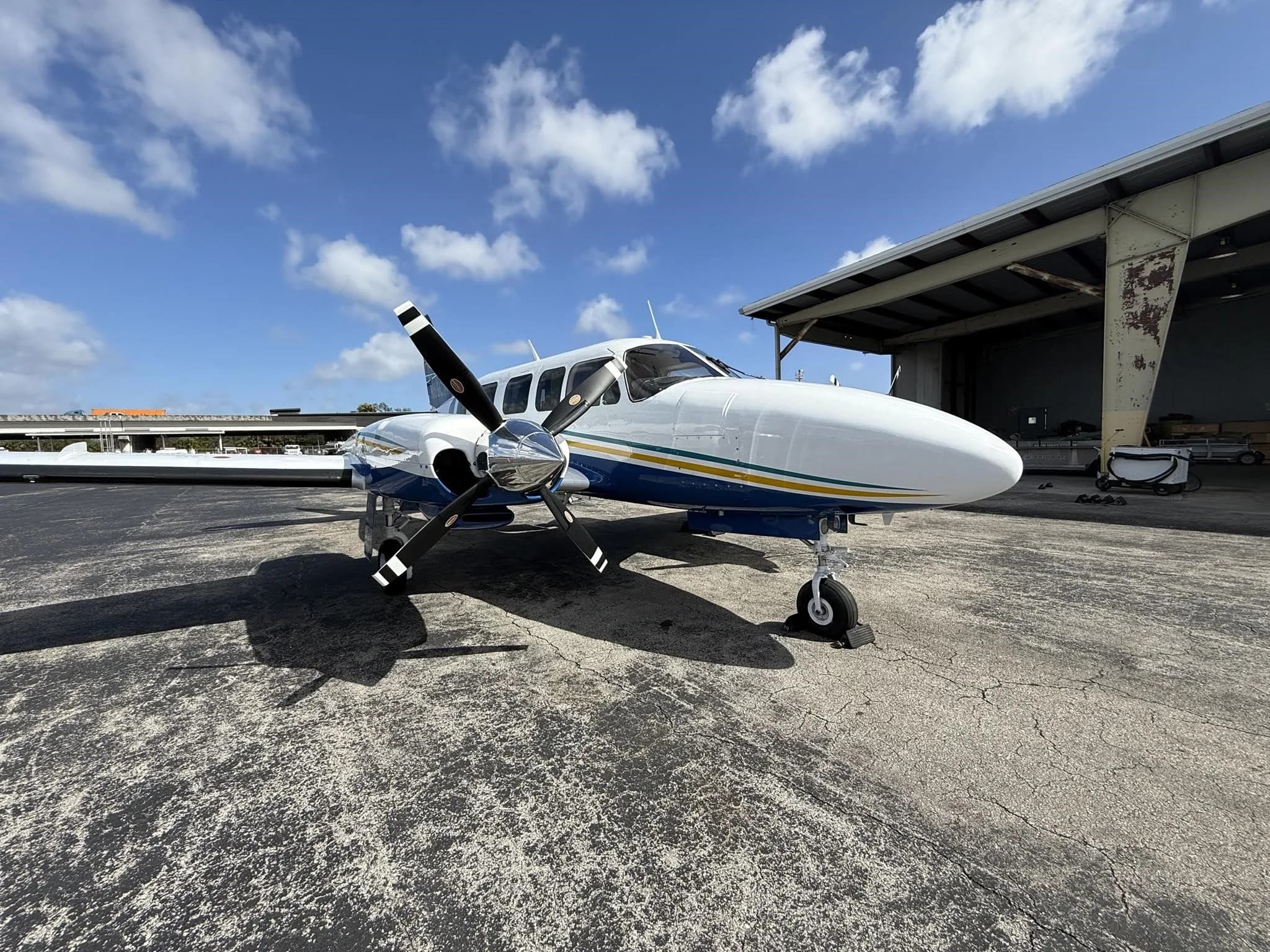 A white and blue twin-engine propeller aircraft with yellow and green stripes, parked on an airport tarmac next to a hangar.