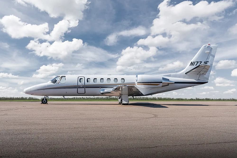 A white Cessna Citation business jet with gold and black stripes is parked on an asphalt tarmac under a partly cloudy sky.