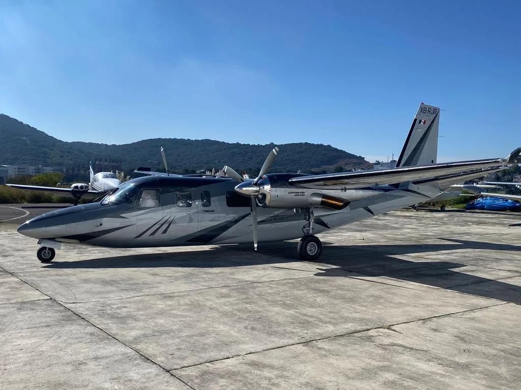 A silver and dark grey Commander 690B twin-engine turboprop, registration XB-RUR, parked on an airfield with mountains in the background.