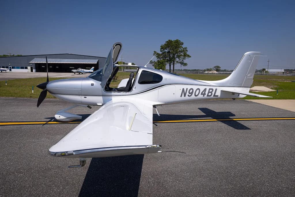 A three-quarter front view of the silver Cirrus SR22 aircraft N904BL with its doors open, parked on an airfield.