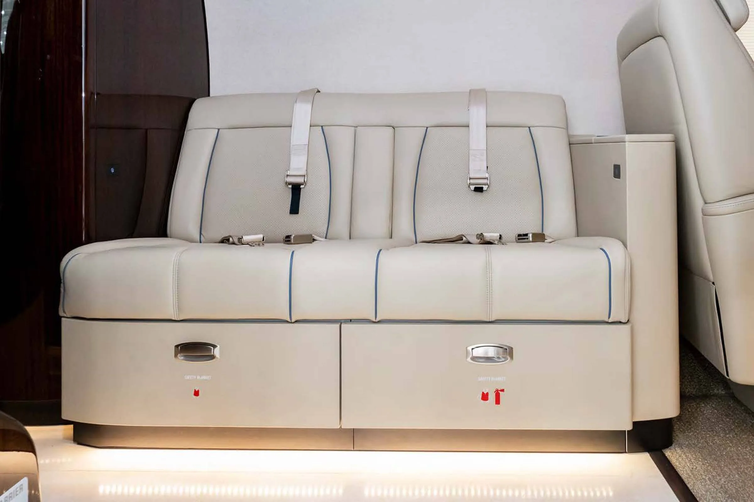 A two-person beige leather bench seat with seatbelts and storage compartments labeled "SAFETY BLANKET" in a private jet cabin.
