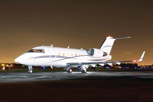 A white business jet with dark stripes is parked on an airfield at night, with airport lights visible in the background.