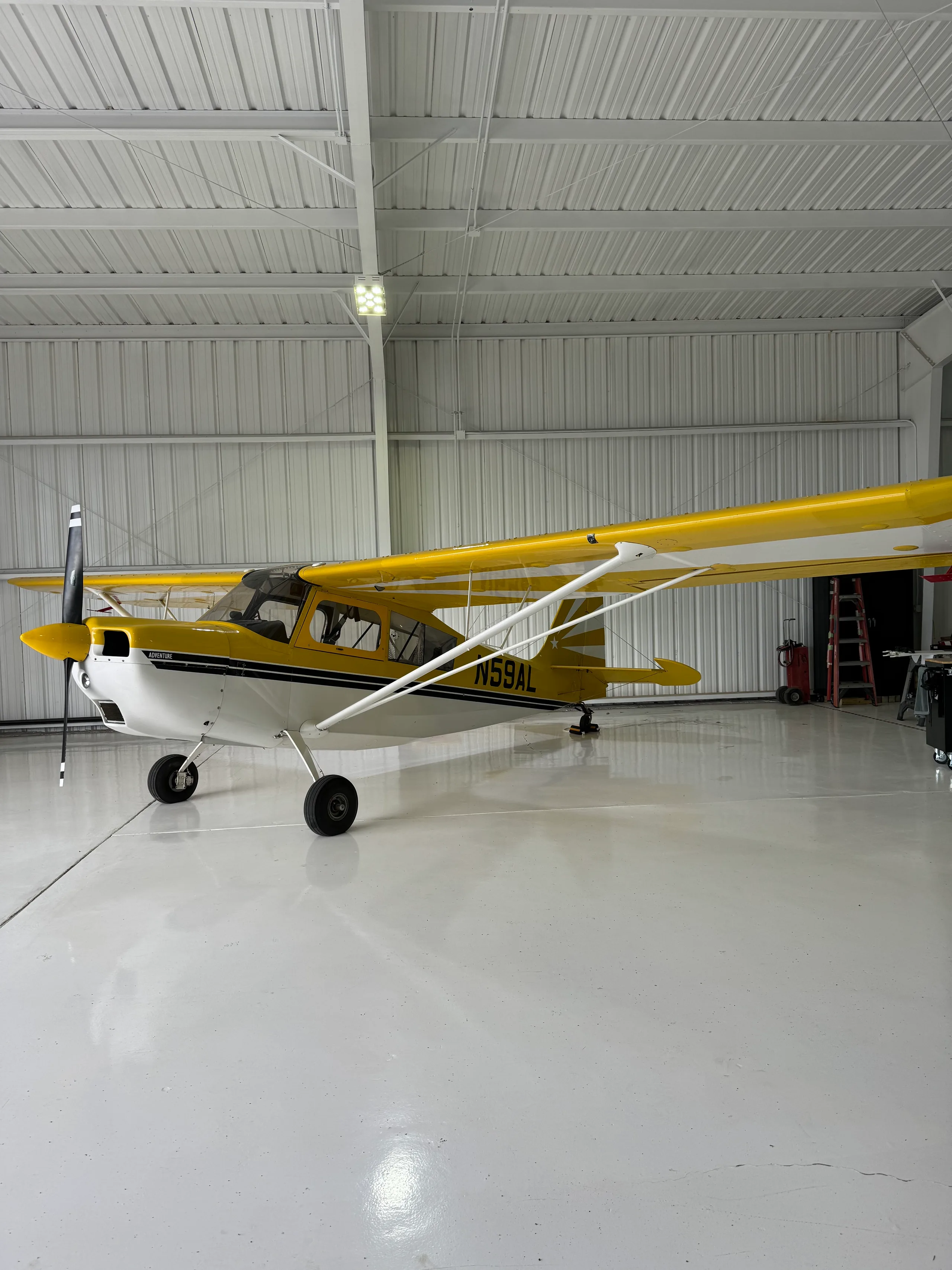 A yellow and white high-wing monoplane, registration N59AL, with 'ADVENTURE' on the cowling, parked inside a brightly lit hangar.